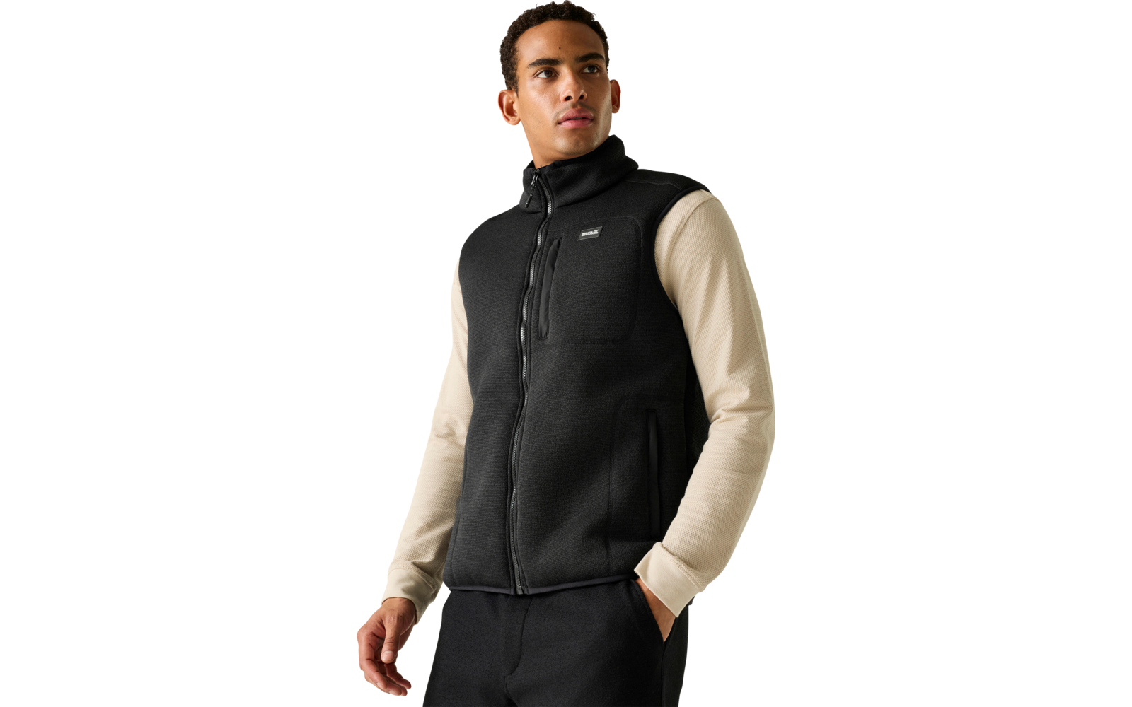 Persoon voor een lichtgrijze achtergrond, draagt een zwarte mouwloze ritssluiting-vest over een beige longsleeve, één hand in de zak, kijkt naar rechts