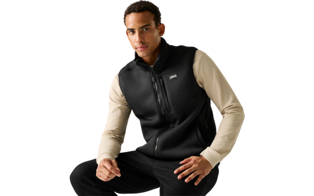 Man zit, draagt een zwarte fleece-vest met rits over een beige shirt met lange mouwen en een zwarte broek, lichte egale achtergrond