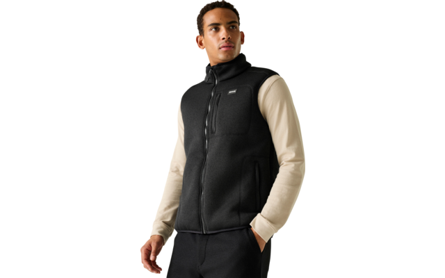 Persoon voor een lichtgrijze achtergrond, draagt een zwarte mouwloze ritssluiting-vest over een beige longsleeve, één hand in de zak, kijkt naar rechts