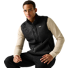 Man zit, draagt een zwarte fleece-vest met rits over een beige shirt met lange mouwen en een zwarte broek, lichte egale achtergrond
