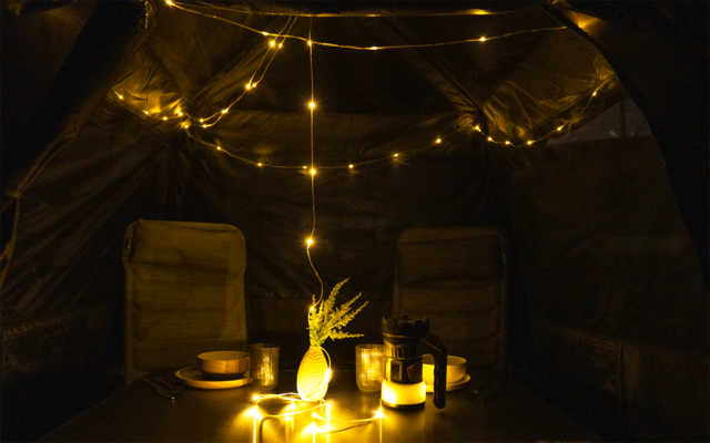 Innenraum eines Zeltes mit Lichterkette an der Decke, zwei Stühlen, einem Tisch mit Teller, Schüssel, Vase mit Zweig und brennenden Kerzen