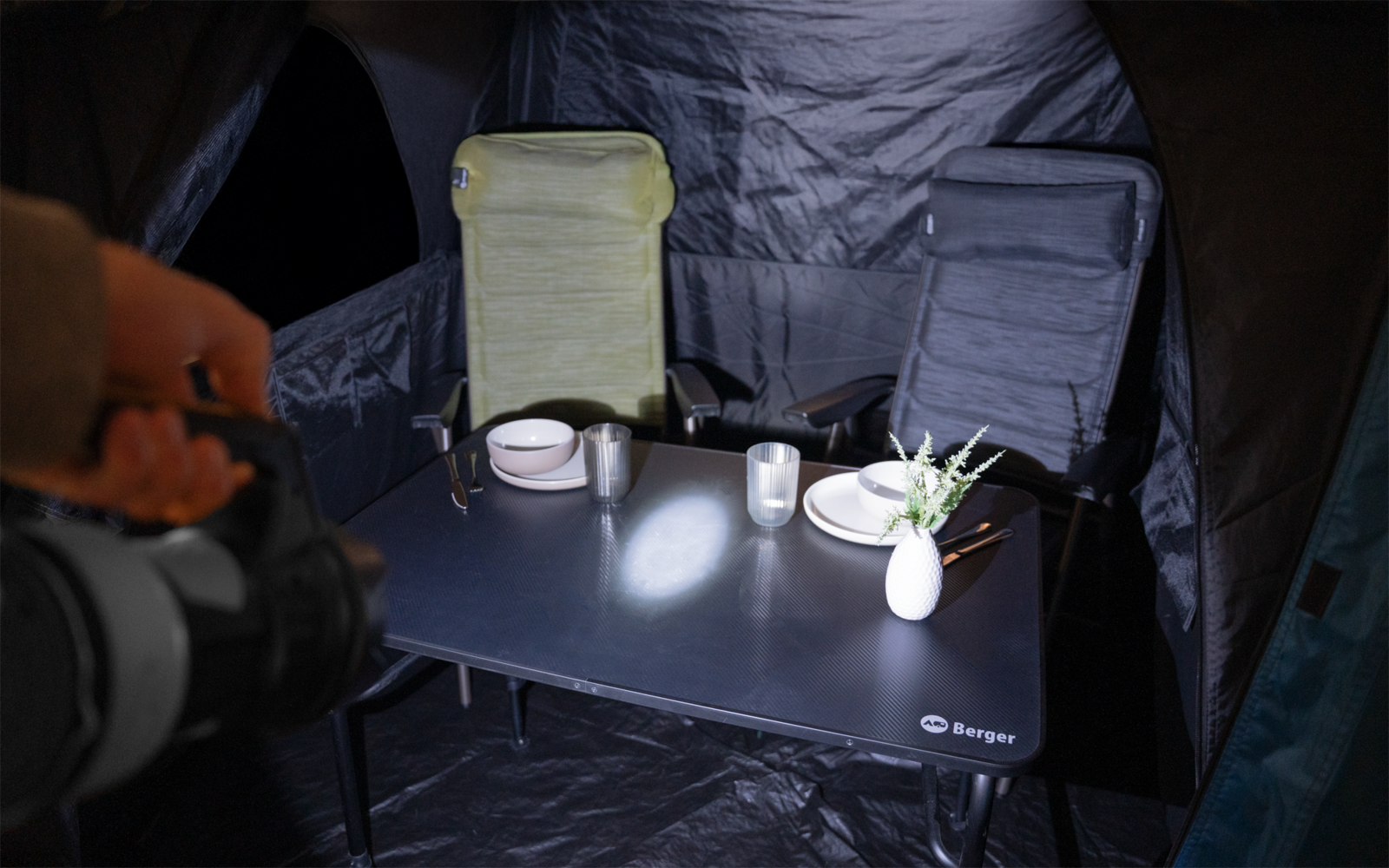 Innenraum eines Zeltes mit Klapptisch, zwei Campingstühlen (ein grüner), Tellern, Tasse, zwei Vasen mit Pflanze und einem Lichtkegel; links Hand mit Kamera sichtbar