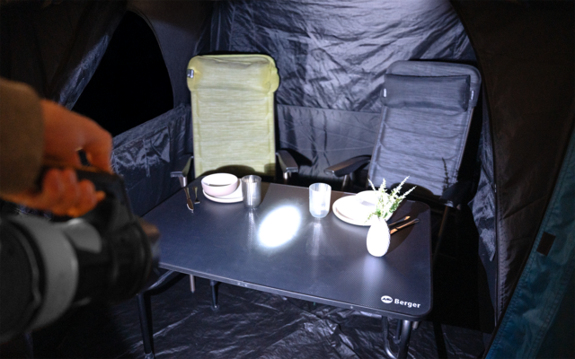 Innenraum eines Zeltes mit zwei Klappstühlen (grün, dunkel), einem schwarzen Tisch mit zwei Tellern, Tassen, einer weißen Vase mit Pflanze und einem Lichtkegel; links eine Hand hält eine Taschenlampe