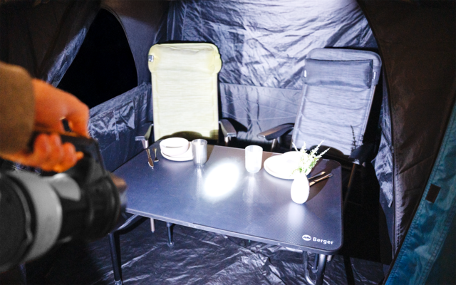 Innenraum eines Zeltes mit Klapptisch und zwei Stühlen. Auf dem Tisch: zwei Becher, eine Schale, eine kleine Topfpflanze und eine Taschenlampe, deren Licht auf den Tisch fällt. Links hält eine Hand die Taschenlampe