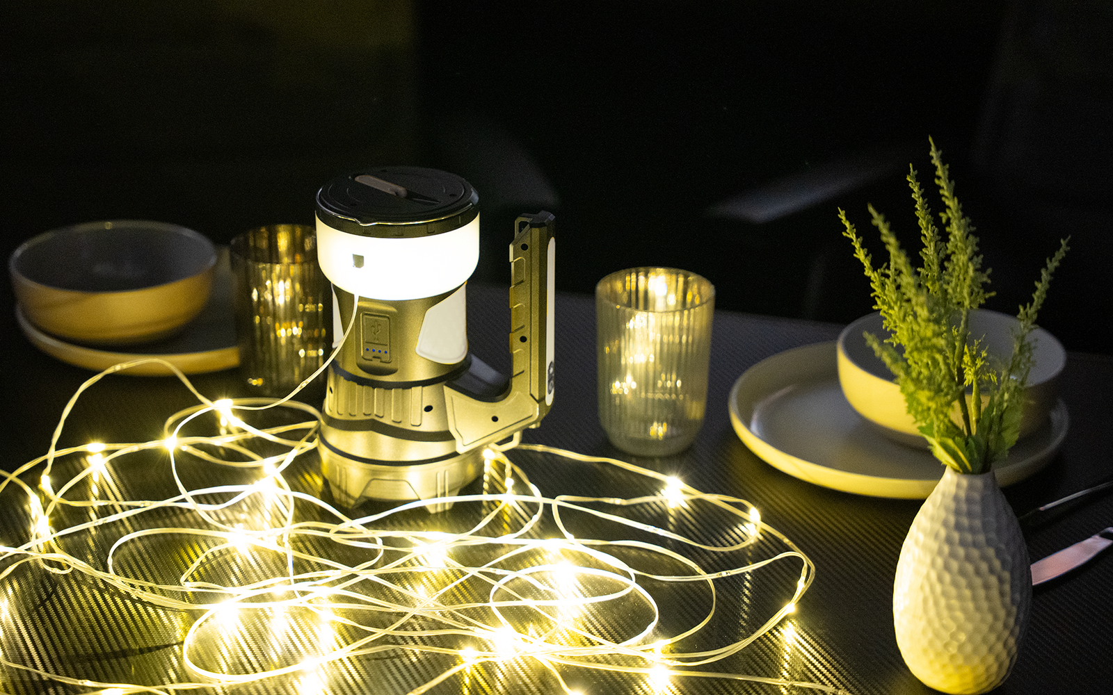 Tisch mit einer Campinglaterne, verstreuten Lichterketten, einem Glaswindlicht mit brennender Kerze, zwei Schüsseln und einer weißen Vase mit grüner Pflanze