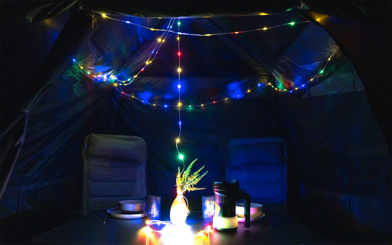 Innenraum eines dunklen Zelts mit bunten Lichterketten an der Decke, zwei Campingstühlen und einem Tisch mit Vase mit grünen Zweigen, Bechern und einer Thermoskanne