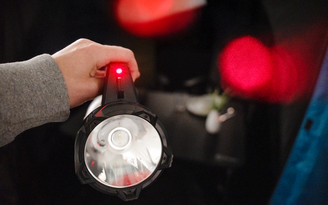 Hand hält Taschenlampe zur Kamera, Lichtkegel sichtbar und rotes Kontrolllicht, unscharfer dunkler Hintergrund