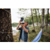 A man stands between trees by a lake holding a camera with a large lens in front of him. A blue hammock to the right, string lights around a tree trunk, buildings on the opposite shore