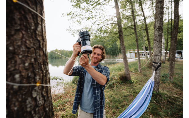 A man stands between trees by a lake holding a camera with a large lens in front of him. A blue hammock to the right, string lights around a tree trunk, buildings on the opposite shore