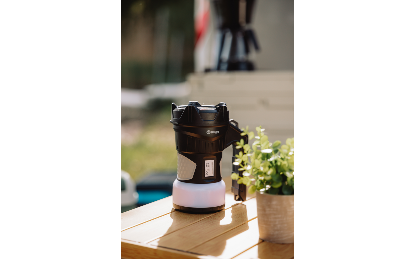 Black portable lantern on a wooden table, next to a small pot with green leaves, blurred background