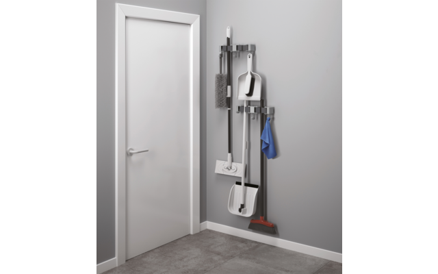 Weiße Tür links; rechts an grauer Wand eine Wandhalterung mit Besen mit roten Borsten, zwei weißen Schaufeln, Mopp, Bürste und blauem Tuch