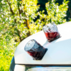 Zwei Gläser mit rotem Getränk stehen auf der weißen Motorhaube eines Autos, im Hintergrund Bäume mit grünen Blättern und gelben Blüten