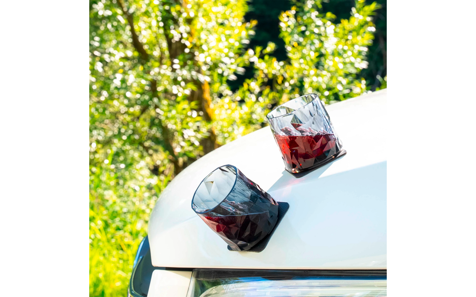 Zwei Gläser mit rotem Getränk stehen auf der weißen Motorhaube eines Autos, im Hintergrund Bäume mit grünen Blättern und gelben Blüten