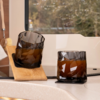 Two faceted amber drinking glasses on a light kitchen countertop by a window; one glass stands on a coaster, the other leans tilted on a small wooden board; a dish brush and blurred trees in the background