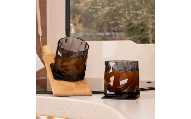 Two faceted amber drinking glasses on a light kitchen countertop by a window; one glass stands on a coaster, the other leans tilted on a small wooden board; a dish brush and blurred trees in the background