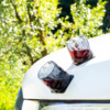 Two faceted glasses with red liquid on a white car hood near a headlight, green foliage in the background