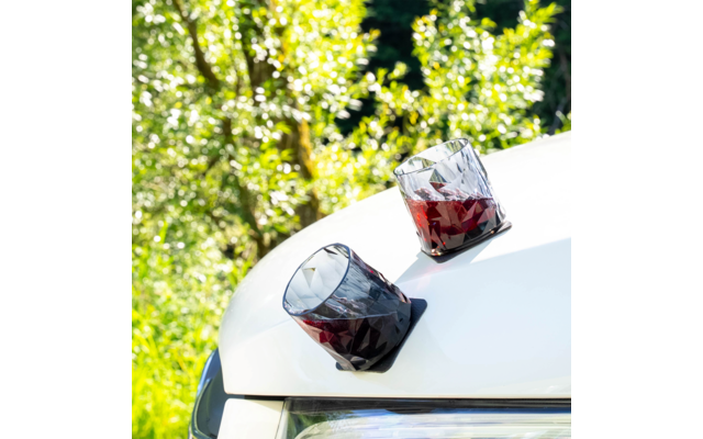 Two faceted glasses with red liquid on a white car hood near a headlight, green foliage in the background