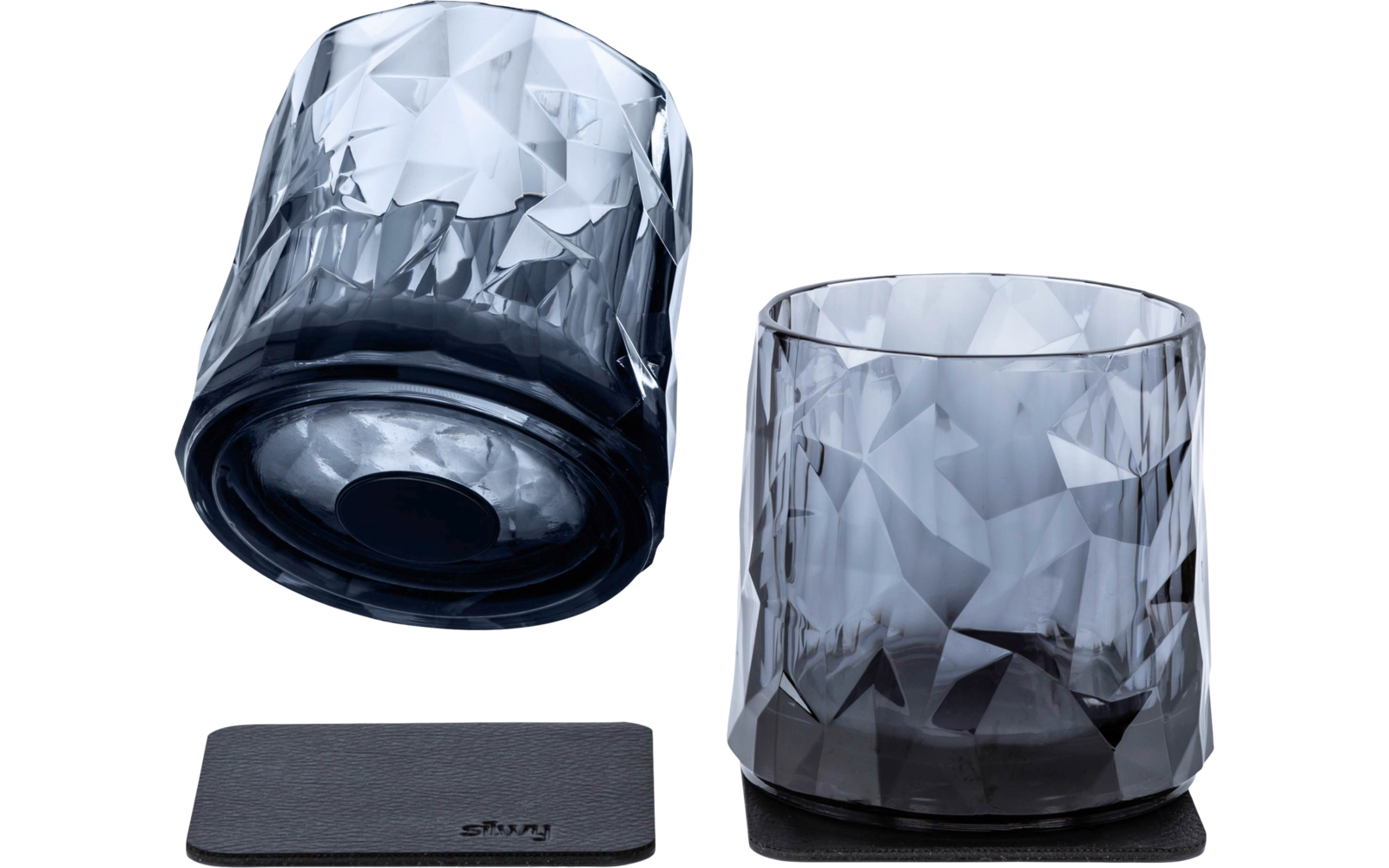 Two short smoky faceted glasses on a white background; one stands on a black square coaster, the other lies on its side on a second coaster