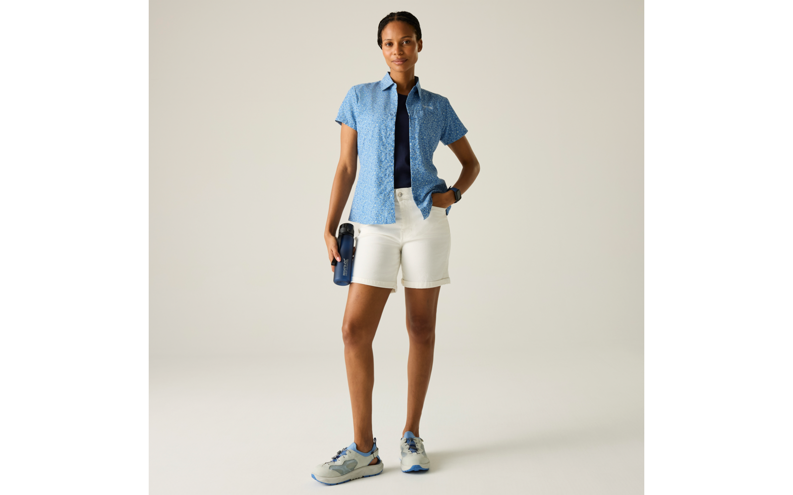 Person steht vor hellem Hintergrund, trägt offenes kurzärmeliges blaues Hemd mit Muster über dunkelblauem T‑Shirt, weiße Shorts, weiß‑blau‑graue Sneakers, hält eine blaue Trinkflasche in der rechten Hand, linke Hand in der Hosentasche