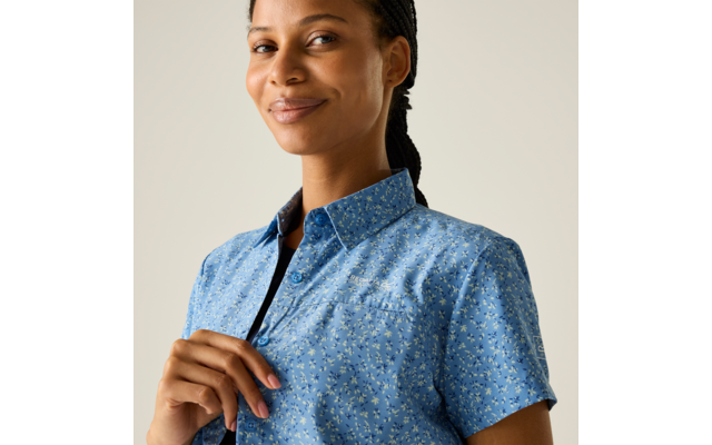 Person mit geflochtenen Haaren, trägt blaues kurzärmliges Hemd mit kleinem Blumenmuster, Hand am Kragen, blickt leicht seitlich