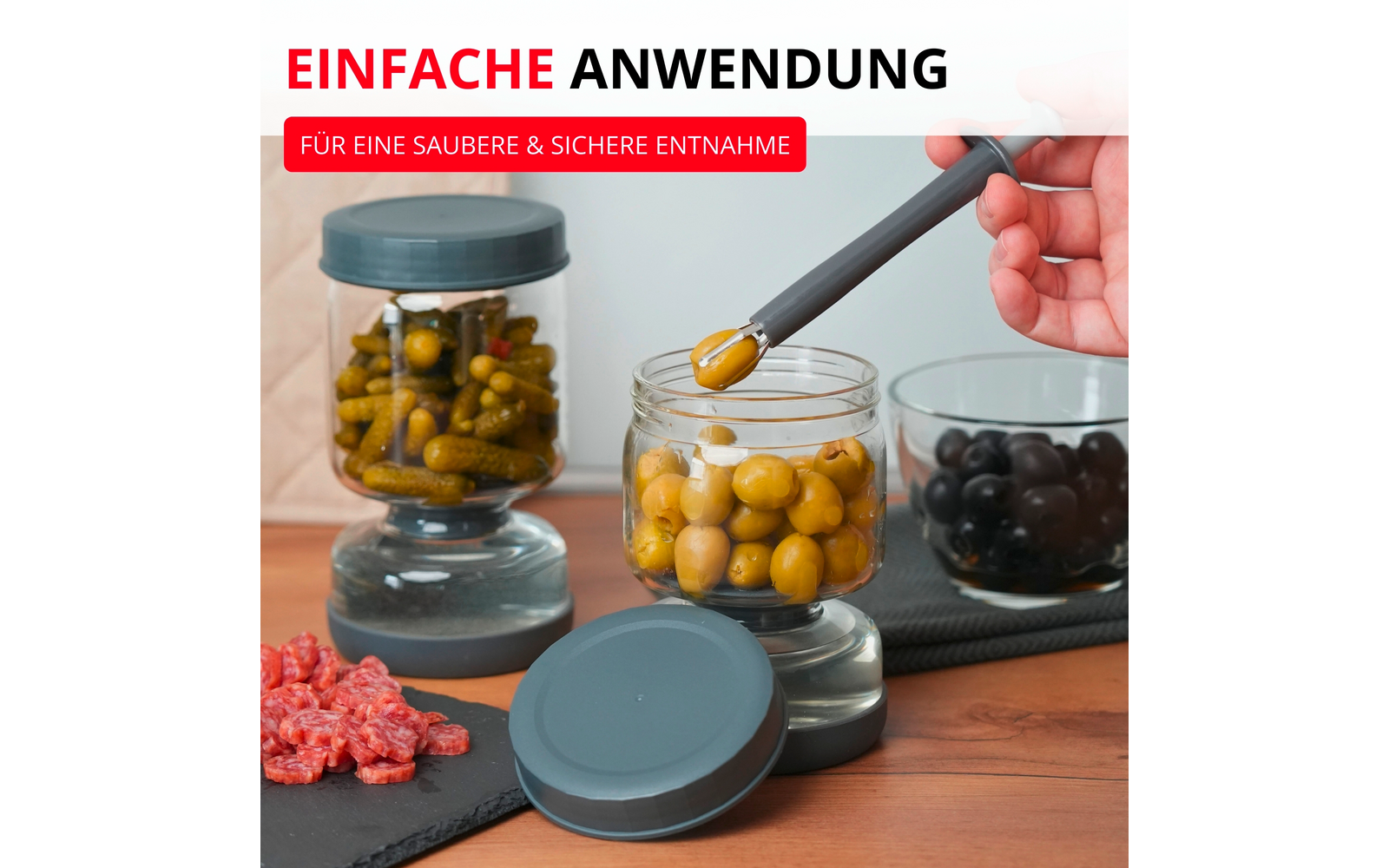 Main tenant une pince grise au‑dessus d'un bocal ouvert d'olives vertes ; deux autres bocaux avec cornichons et olives noires sur socles en verre, couvercle gris sur surface en bois ; bandeaux rouges en haut indiquant 'EINFACHE ANWENDUNG' et 'FÜR EINE SAUBERE & SICHERE ENTNAHME'