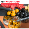 Outil métallique à deux branches tient une olive jaune au-dessus d'un bocal d'olives jaunes; olives noires et dés de poivron rouge en arrière-plan; en haut texte KEIN WEGRUTSCHEN et bandeau rouge EDELSTAHLKRALLE GREIFT ALLES BIS 3 CM Ø