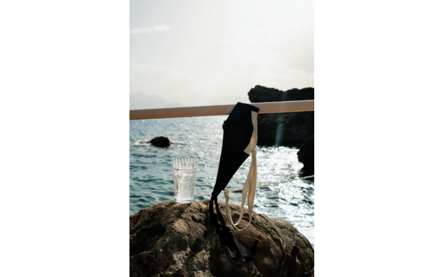Haut de bikini noir aux bretelles blanches accroché à une rambarde blanche; un verre d'eau posé sur un rocher; mer et côte rocheuse en arrière-plan
