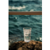 Verre transparent avec eau pétillante sur un rocher, mer floue en arrière-plan et une rambarde horizontale au-dessus