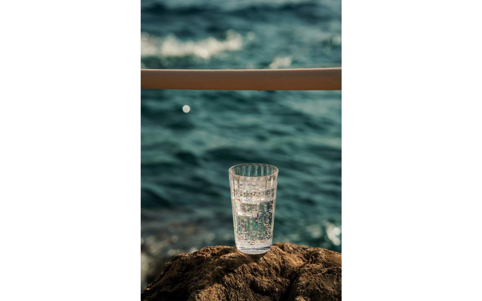 Verre transparent avec eau pétillante sur un rocher, mer floue en arrière-plan et une rambarde horizontale au-dessus
