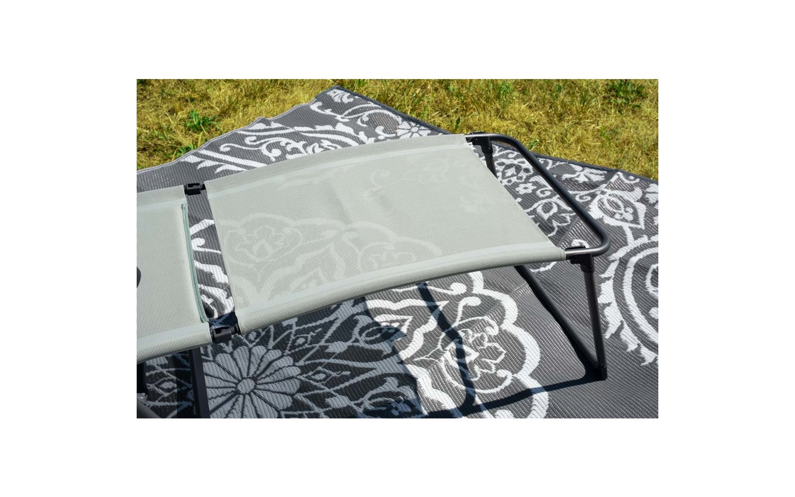 Teil eines hellgrauen Stoff-Klappstuhls mit schwarzem Metallrahmen und Riemen auf einer schwarz-weißen, gemusterten Outdoor-Matte; oben Rasen