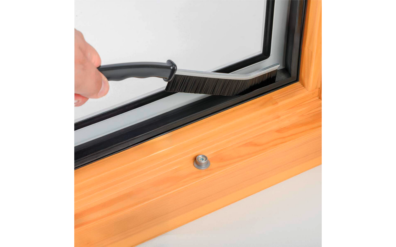 Mano sosteniendo un cepillo pequeño junto al borde interior de una ventana de doble acristalamiento con marco de madera; sujeción metálica en el alféizar
