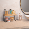 Kleine Holzablage mit schwarzen Metallfüßen auf einer Arbeitsfläche; auf der Ablage ein Metallbecher mit Make-up-Pinseln und zwei rechteckige Glasflaschen; unter der Ablage drei kleine Cremedöschen; rechts eine Glasflasche mit dünnen Stäbchen; Rand e