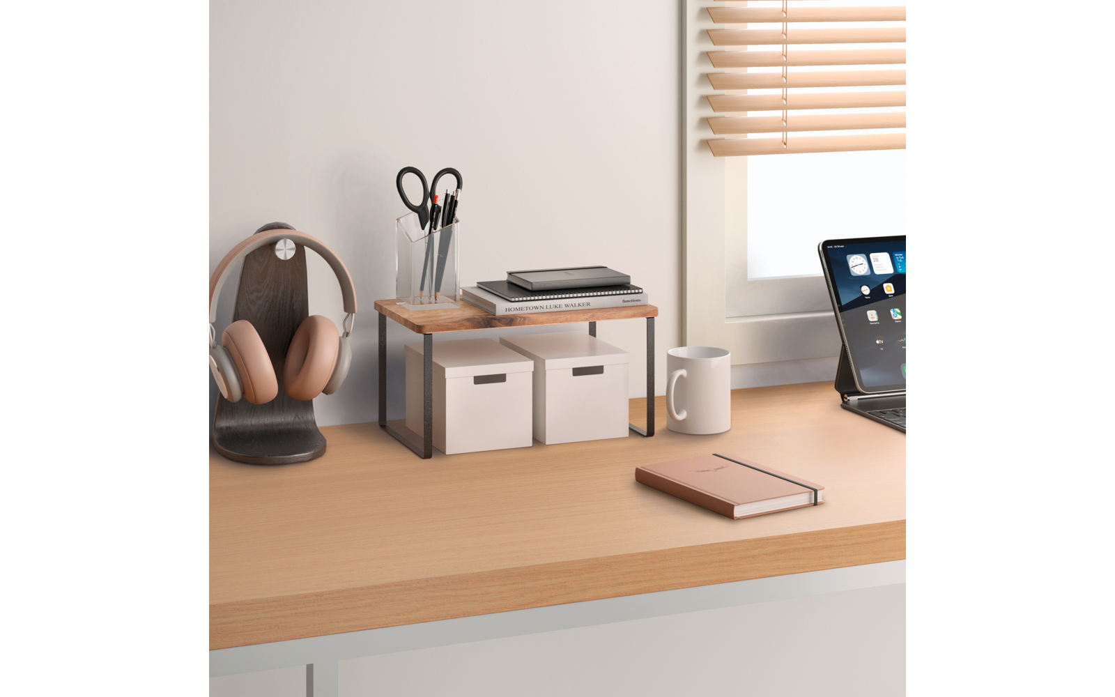 Schreibtisch mit Kopfhörern auf einem Ständer, kleinem Holzregal mit zwei weißen Aufbewahrungsboxen und Stapel Notizbücher, Stiftebehälter mit Schere und Stiften, Kaffeetasse, geschlossenes Notizbuch, Tablet mit Display und Fenster mit Jalousien