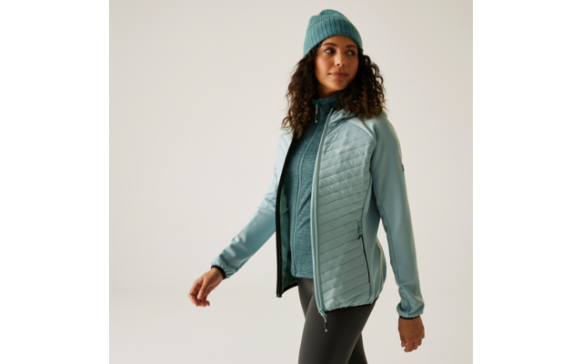 Femme aux cheveux bouclés portant une veste bleue, un pull bleu et un bonnet turquoise devant un fond blanc