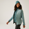 Femme aux cheveux bouclés portant une veste bleue, un pull bleu et un bonnet turquoise devant un fond blanc