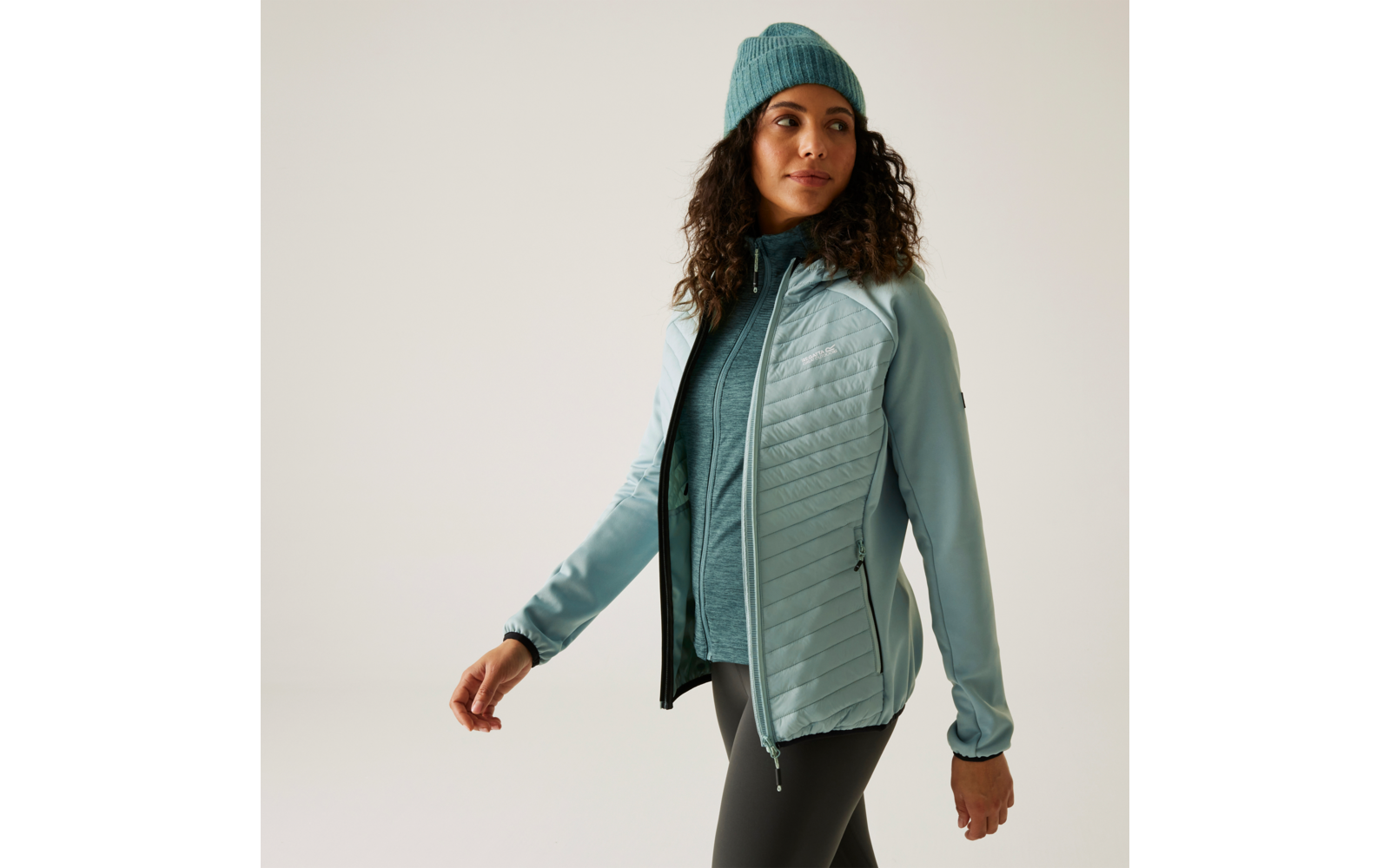 Femme aux cheveux bouclés portant une veste bleue, un pull bleu et un bonnet turquoise devant un fond blanc