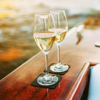 Zwei Weingläser mit weißem Wein stehen auf einem Holzgeländer mit Wasser im Hintergrund