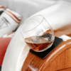 Verre rond contenant un liquide ambré posé sur un sous-verre noir sur un accoudoir en bois ; housse blanche et coussin rayé en arrière-plan