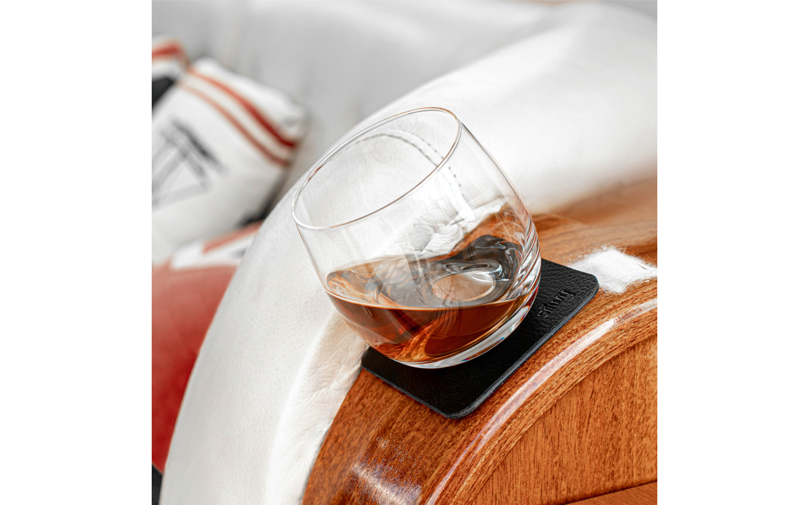 Verre rond contenant un liquide ambré posé sur un sous-verre noir sur un accoudoir en bois ; housse blanche et coussin rayé en arrière-plan