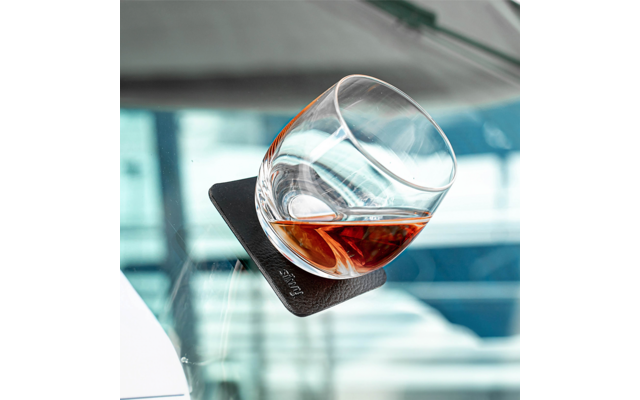 Verre rond incliné contenant un liquide brun sur un dessous-de-verre carré noir posé sur une surface vitrée ; arrière-plan bleu flou