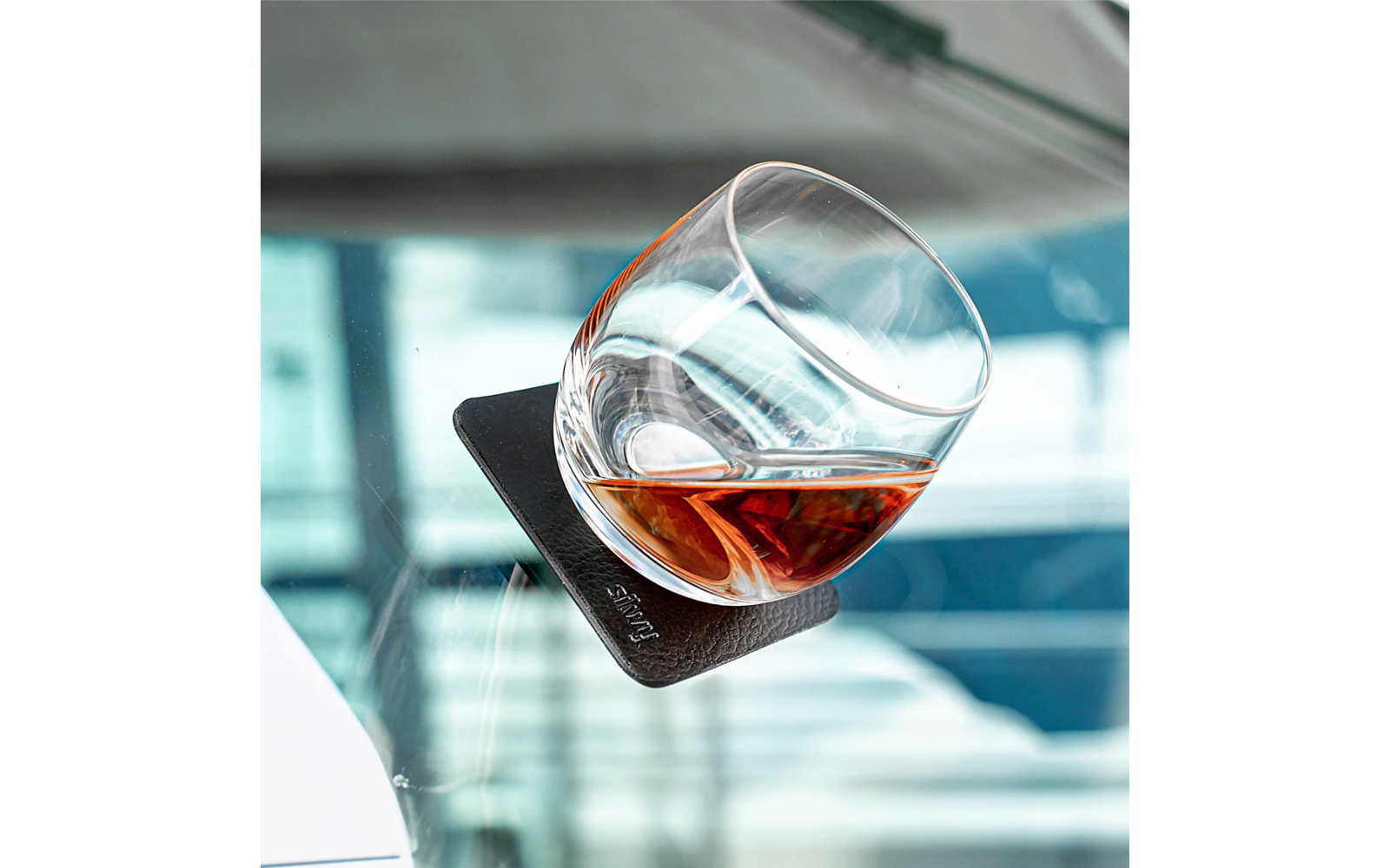 Verre rond incliné contenant un liquide brun sur un dessous-de-verre carré noir posé sur une surface vitrée ; arrière-plan bleu flou