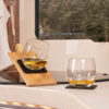 Deux verres sans pied avec un liquide ambré sur un plan de travail clair près d'une fenêtre ; un verre est incliné dans un support en bois, l'autre repose sur un sous-verre foncé ; à gauche, une brosse et une serviette pendent