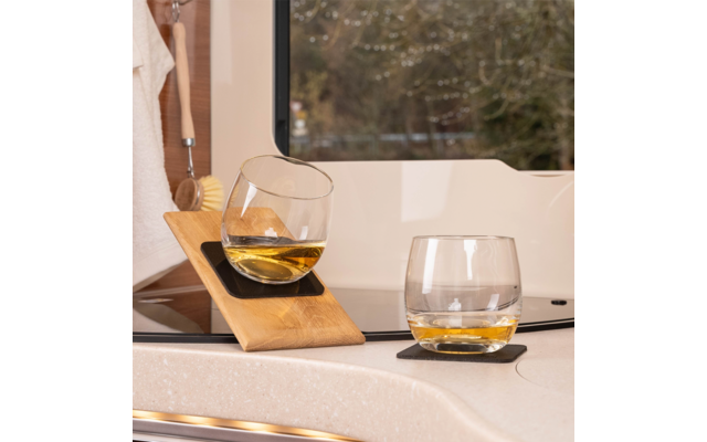 Deux verres sans pied avec un liquide ambré sur un plan de travail clair près d'une fenêtre ; un verre est incliné dans un support en bois, l'autre repose sur un sous-verre foncé ; à gauche, une brosse et une serviette pendent