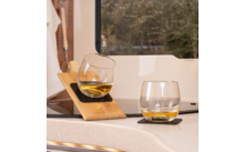 Two stemless glasses with amber liquid on a light countertop by a window; one glass is tilted in a wooden holder, the other stands on a dark coaster; a brush and towel hang to the left
