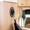 Holzverkleidung mit rundem schwarzem Wandhalter und Metallhaken, an dem eine schwarze Jacke hängt; im Hintergrund verschwommener Sitz und Fenster