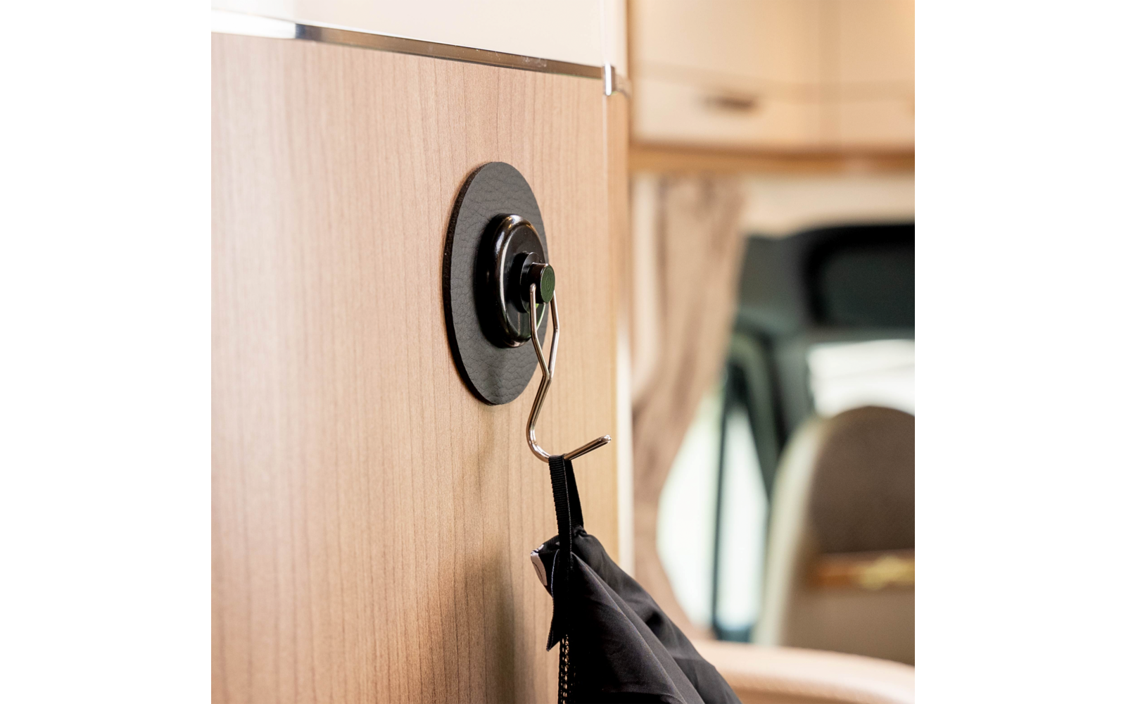 Holzverkleidung mit rundem schwarzem Wandhalter und Metallhaken, an dem eine schwarze Jacke hängt; im Hintergrund verschwommener Sitz und Fenster