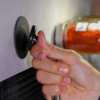 Hand hält eine kleine runde schwarze Saugnapf-Vorrichtung mit Metallstange an einer hellen Wand; unscharfes Glasgefäß mit roten Inhalten im Hintergrund