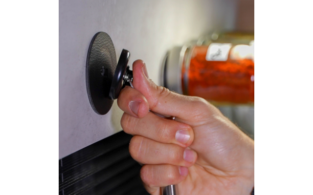 Hand hält eine kleine runde schwarze Saugnapf-Vorrichtung mit Metallstange an einer hellen Wand; unscharfes Glasgefäß mit roten Inhalten im Hintergrund