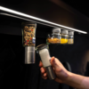 Hand removes a spice jar filled with coarse salt from a row of spice jars mounted on a rail under a light strip; three other jars with colorful spices visible