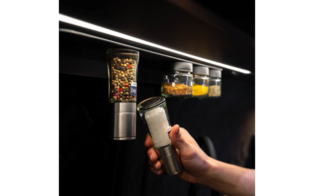 Hand removes a spice jar filled with coarse salt from a row of spice jars mounted on a rail under a light strip; three other jars with colorful spices visible